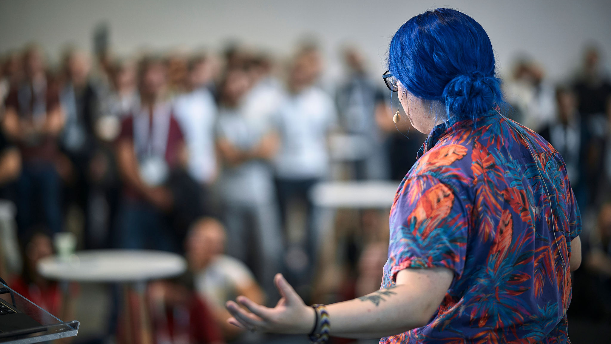 A photo, shot from behind, of a person with blue hair and a colorful shirt presenting to an audience. The audience in the background is blurry.