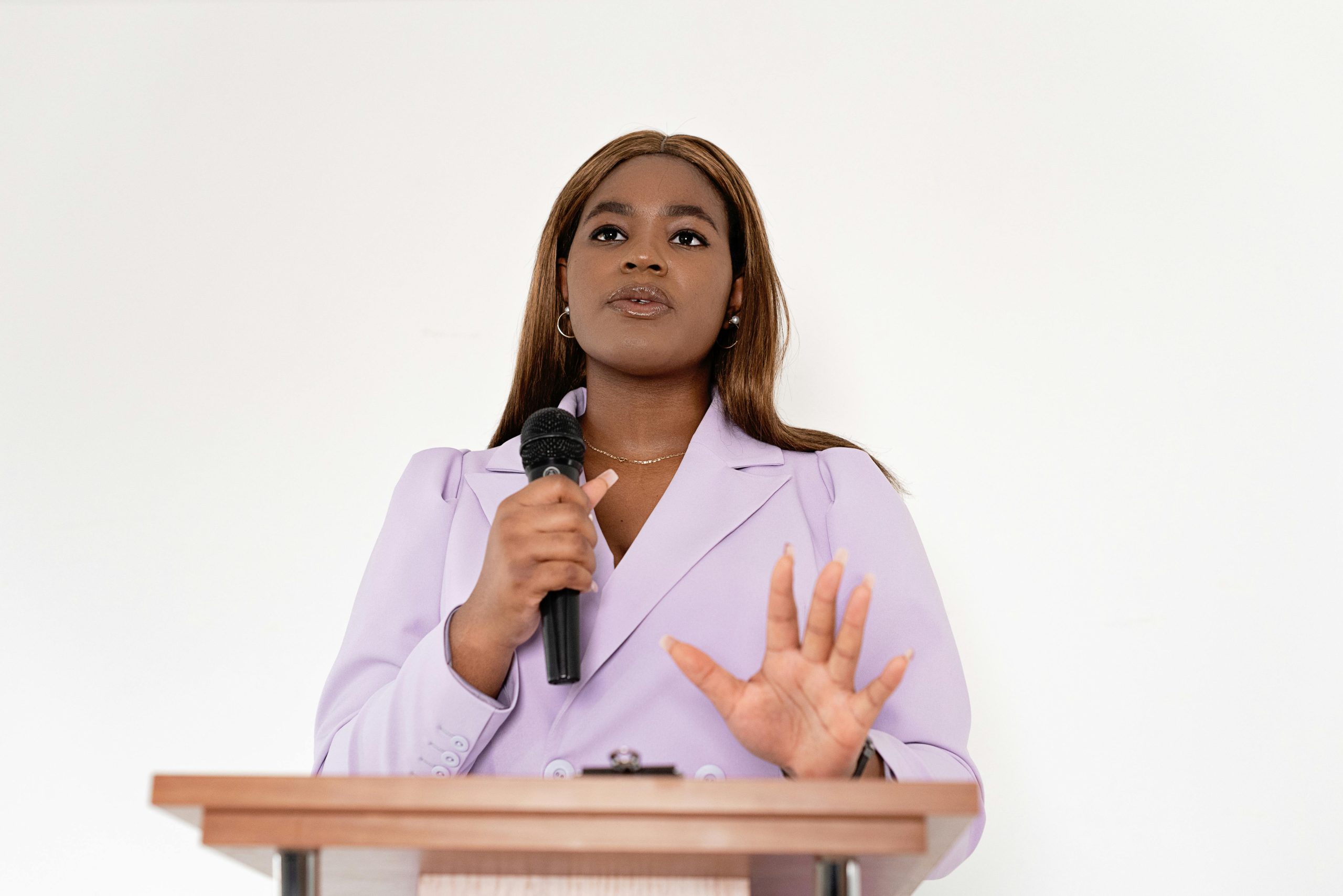 A black woman dressed in formal clothing is standing on front of a podium, holding a microphone. She is speaking and gesturing.